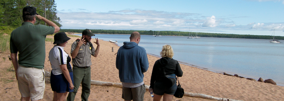 A Park Ranger talking to a group of four visitors on a sandy beach looking out on a lake with sailboats.