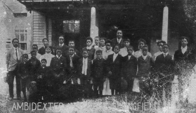 A black and white photograph of teachers standing in front of a wooden house.