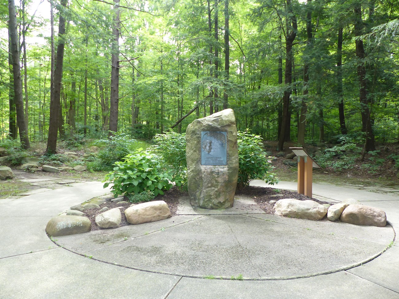 A small circular plaza surrounded by woods. In the center is an upright boulder with a bronze plaque. To the right is an interpretive panel in profile. Landscaping includes bushes, large rocks, and cement pavers.