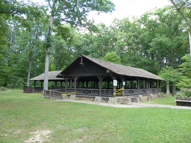 A large T-shaped shelter stands in a wooded picnic area. The open building is made of rough-cut logs with a sandstone foundation and a shingled roof. Yellow caution tape blocks the entrance steps and ramp.