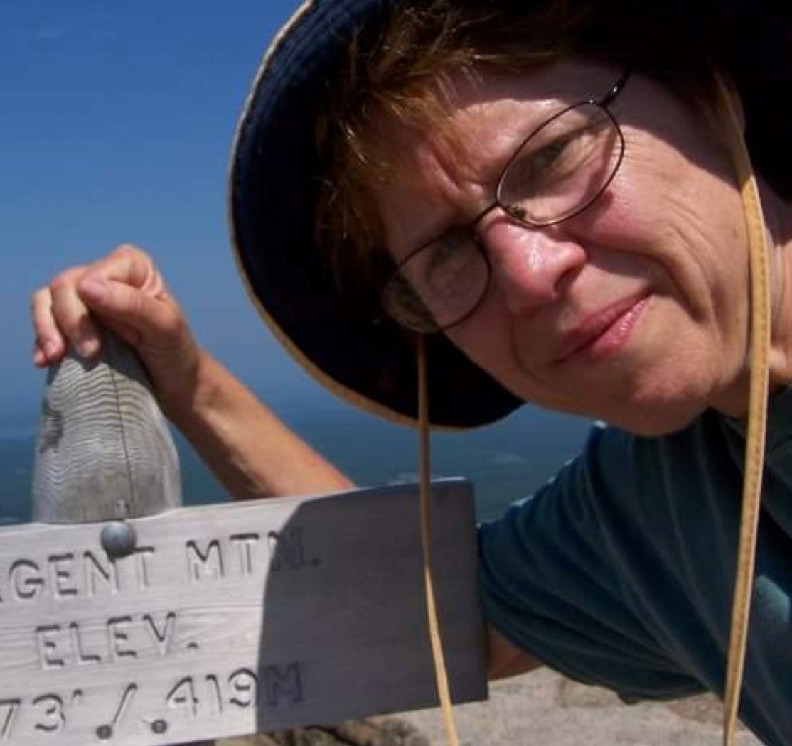 A woman in a sunhat standing next to a wooden sign. The text on the sign is partially cut off, but designates the mountain peak's name and elevation.