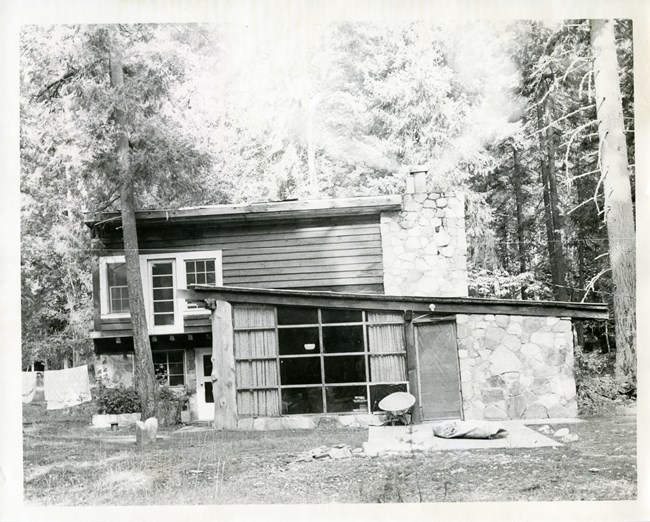 A black and white photo of a flat-roofed multi-room house with wooden paneling and stone work, set in a sparse forest.