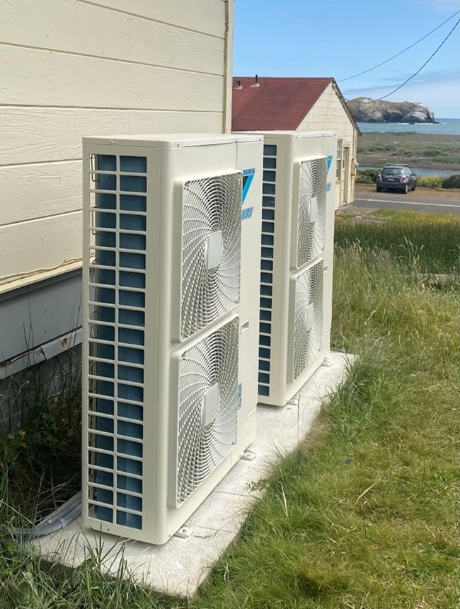 Two heat pumps sit on a pad outside a building at Fort Cronkhite, with the ocean in the background.