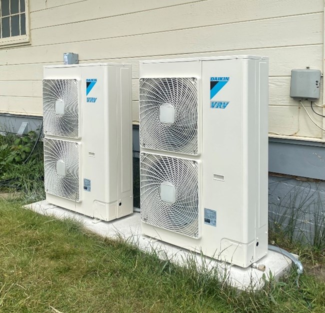 Two heat pumps sit on a pad outside a building at Fort Cronkhite