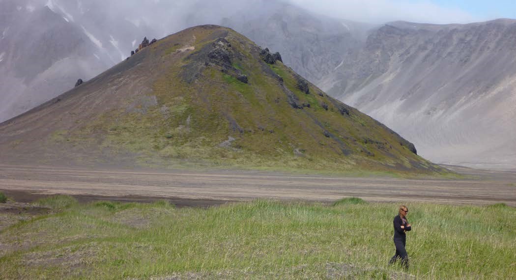 a person walking past a dome-shaped hill with a portion of the volcanic crater rim showing in the distance