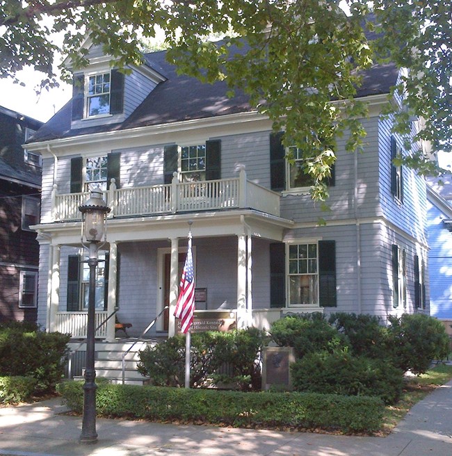 A light gray house with dark green shutters. A flag pole and lamp post are at the front. Small shrubs line the edge of the house.