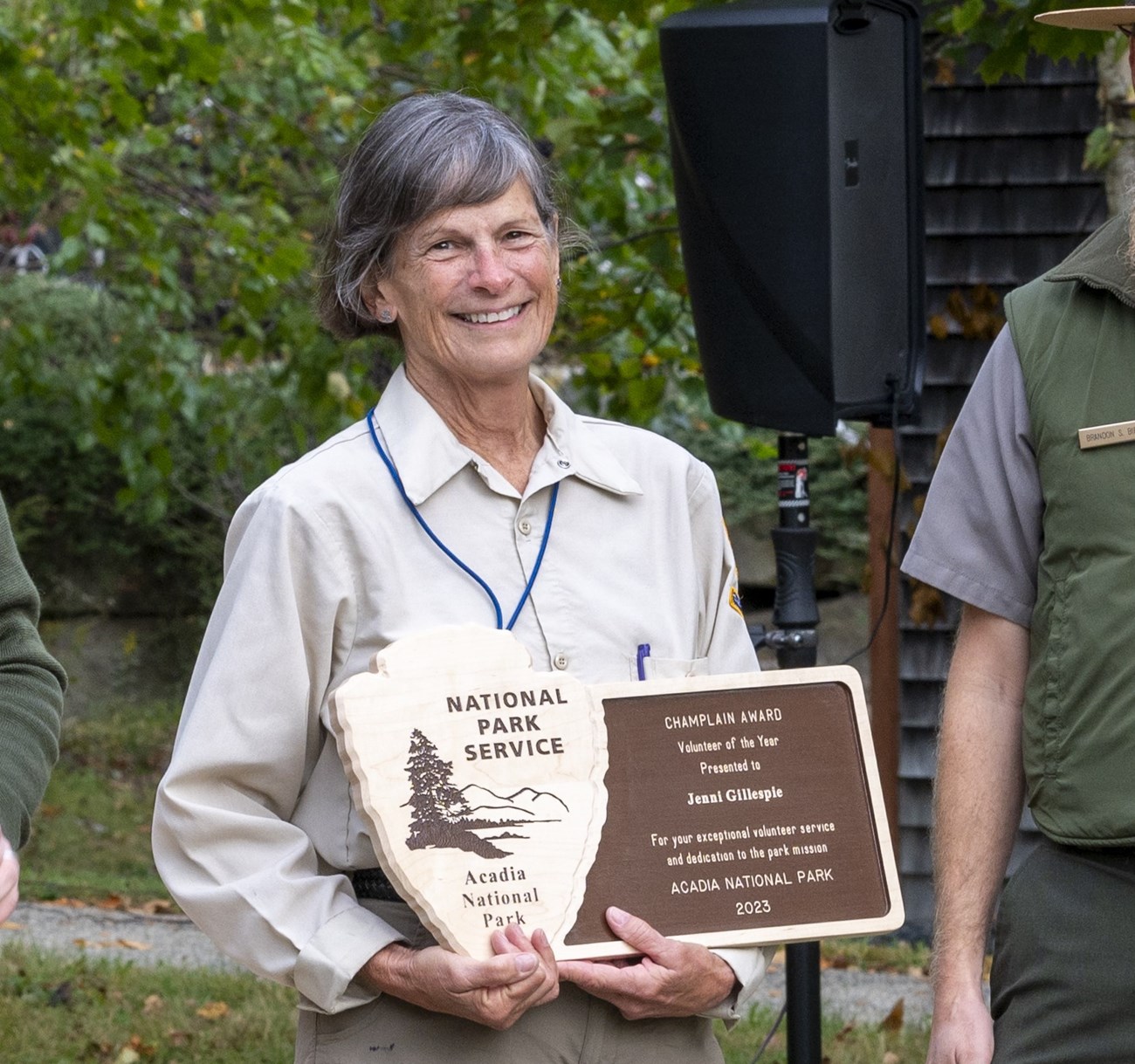 A woman with salt and pepper hair and dressed in the NPS volunteer uniform holds a wooden plaque decorated with the National Park arrowhead logo and the text "Volunteer of the Year: 2023"