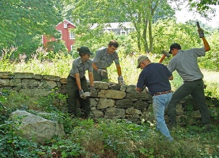 A park ranger places a rock on a stone wall while three stone masons work on the wall alongside her.
