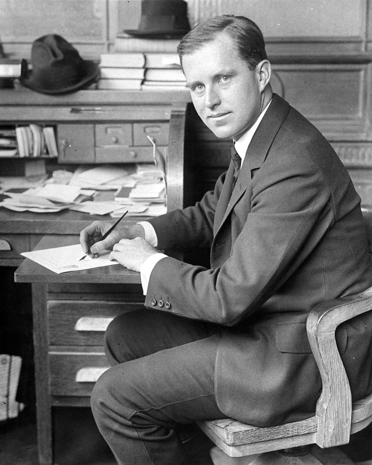 Joseph P. Kennedy sits at a desk holding a pen.