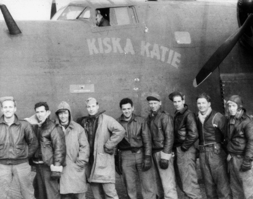 Men in uniform stand in front of plane that has "Kiska Katie" painted on it.