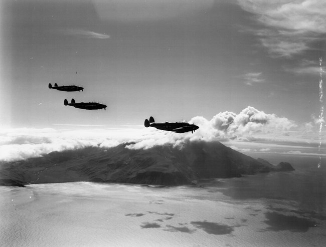 Three planes fly in front of a cloud-capped volcano, with the ocean below.