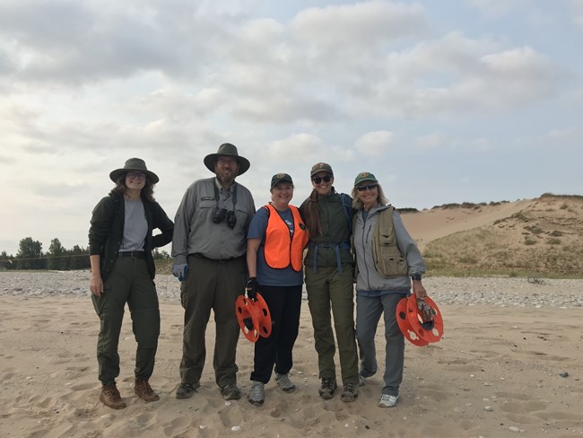 A group of five people stand on a beach. Three are NPS employees wearing the uniform. Two are NPS volunteers.