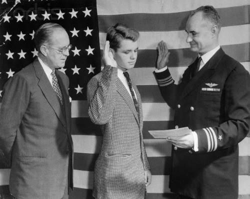 A black and white photo of Bobby Kennedy with his hand raised.  He stands beside his father and a U.S. Navy Officer in front of an American flag.
