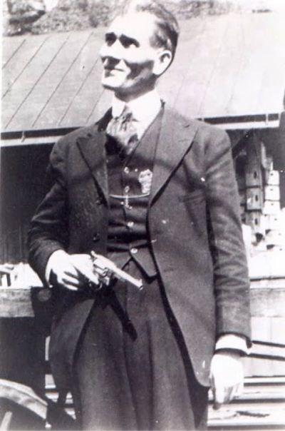 A man in a formal suit with swept back hair stands erect with his head tilted upwards towards the sky and a revolver in his right hand.