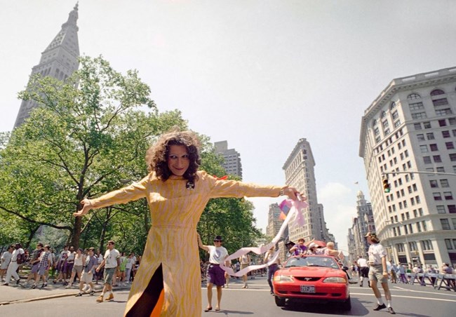 Sylvia Rivera is standing in the streets of NYC in a yellow dress in front of a red car