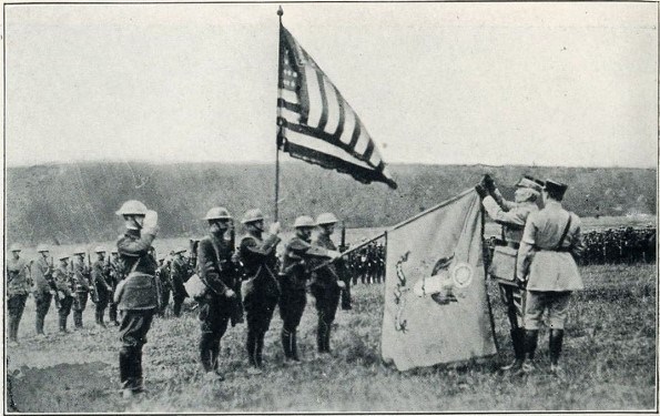 Black and white photo of soldiers standing at attention under an American flag. Two uniformed men are pinning a medal atop a National Guard flag.