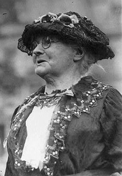 An elderly woman wearing glasses with a black cloth hat and a black and white dress.