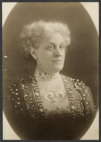 Formal portrait, head and shoulders, Carrie Chapman Catt, facing forward with head and eyes turned slightly to right, wearing a high-collared, richly brocaded or embroidered dress.