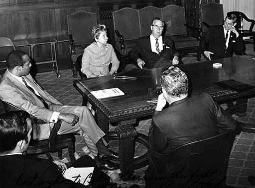 A black-and-white photo shows Betty Klaric, the mayor, and four other men in suits sitting around a large wooden conference table.