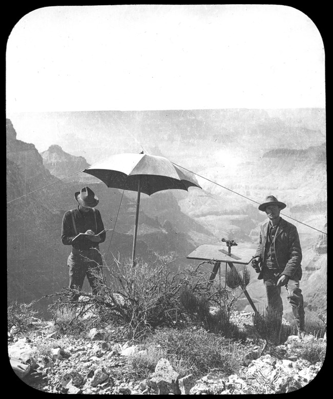 Two men standing with a navigation device and an umbrella between them. Grand Canyon backdrop.
