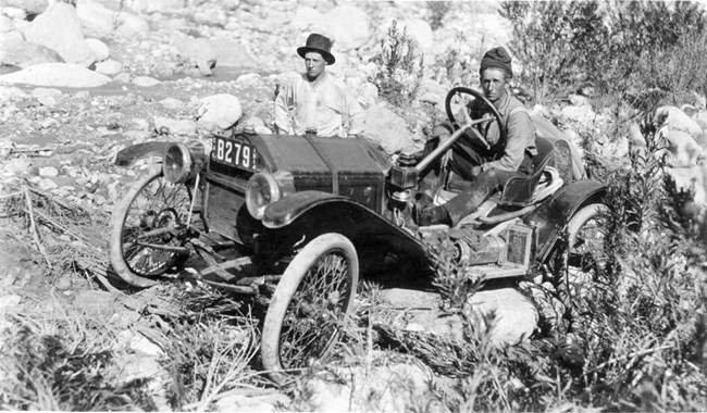 Car among rocks and shrubs with two people inside.