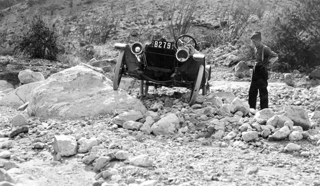 Car stands on a rock pile at an uneven angle, facing front, while the driver stands to the side.