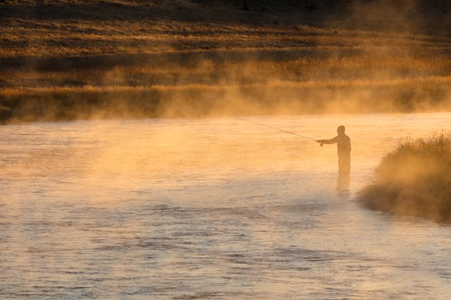 An angler fly fishes in a river at sunrise as fog rises from the water.