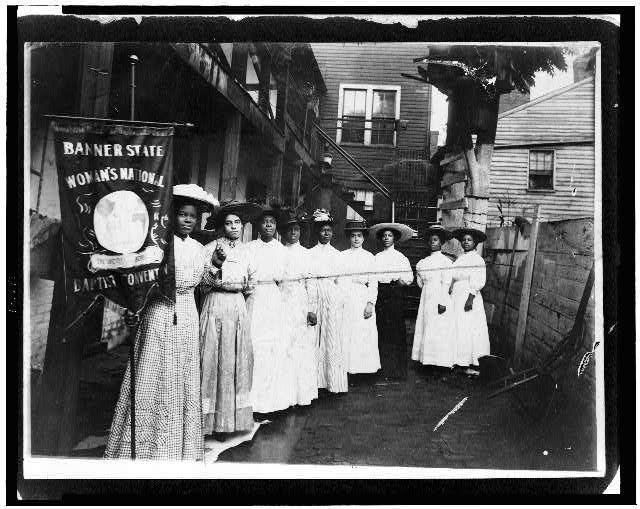 Nine Afro-American women posed, standing, full length, with Nannie Burroughs holding banner reading, "Banner State Woman's National Baptist Convention"