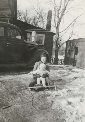 Two children on a sled in the snow with a car parked behind them.