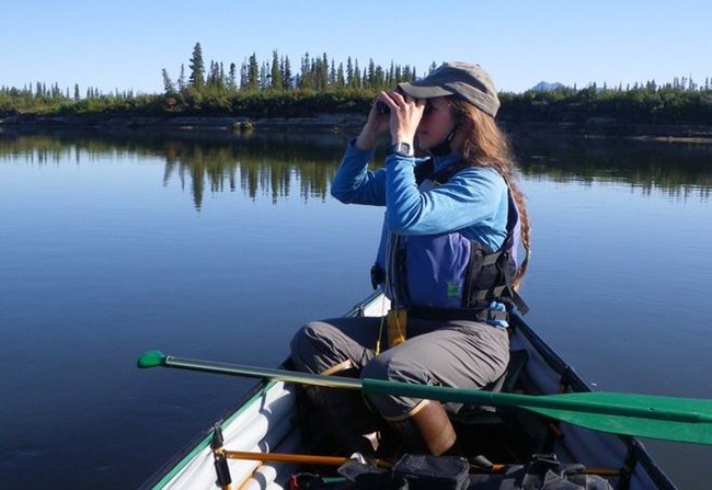 A woman looks through binoculars while sitting in a canoe on a lake