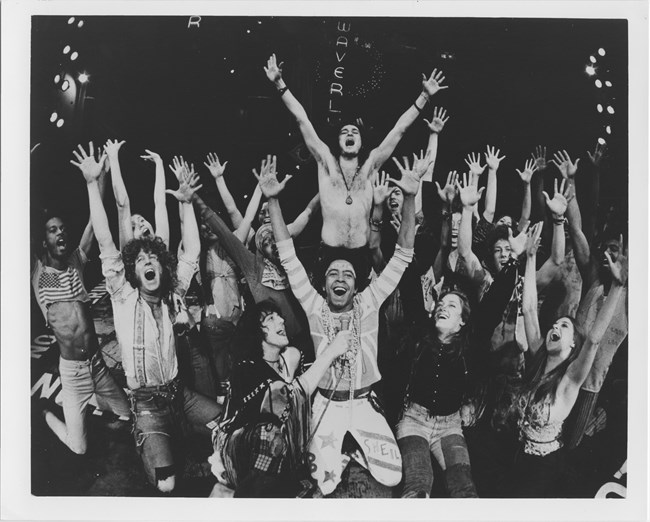 The cast of "Hair" throw their arms in the air, singing.