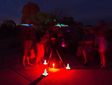 a tripod is illuminated with red light at its base as shapes of people move around it