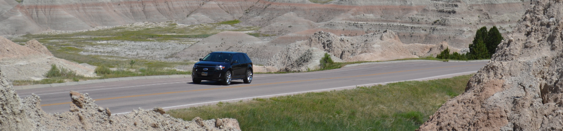 a black suv drives up a grassy hill surrounded by brown badlands buttes.