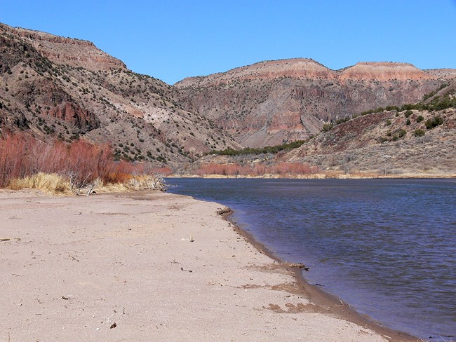 a wide blue river with a sandy beach and tall pinkish cliffs