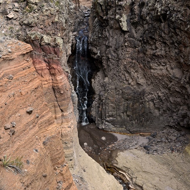 a waterfall drops over black rock surrounded by layers of reddish rock