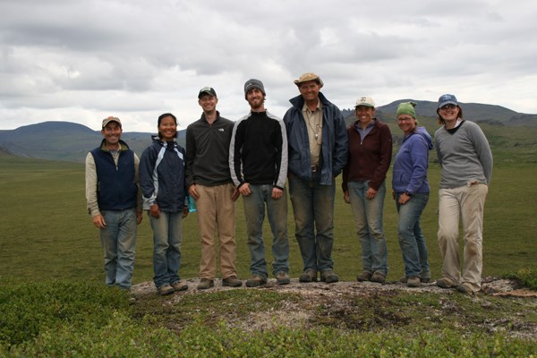 An excavation crew of 8 men and women stands in a row on the tundra, smiling.