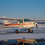 A small white and orange high-winged, single engine aircraft sitting on an airstrip with snow and its reflection in melt water.