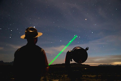 A ranger stands next to a telescope, with a laser pointer shooting towards a stat.