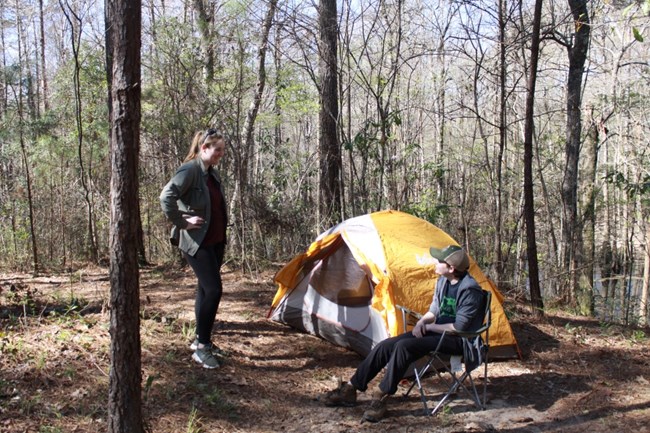 1 person standing and 1 person sitting outside of a tent in the woods