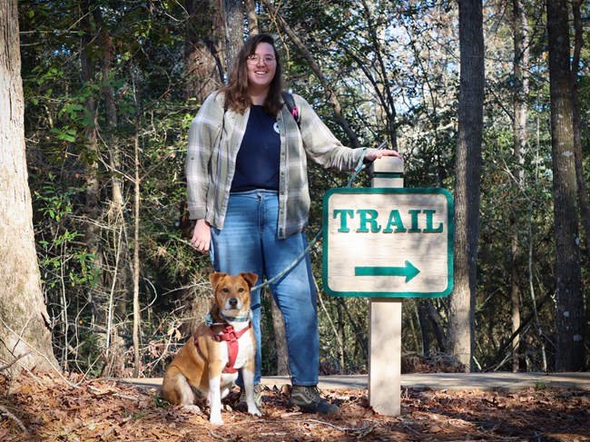 a person standing with a brown and white dog on leash next to a TRAIL sign with an arrow.