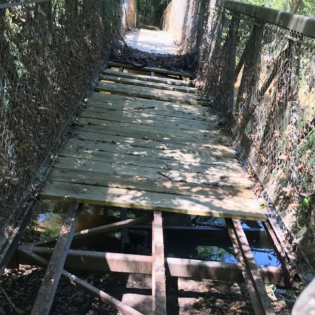 photo of a bridge showing many missing wooden boards, exposing the iron framework beneath and a creek visible below that.