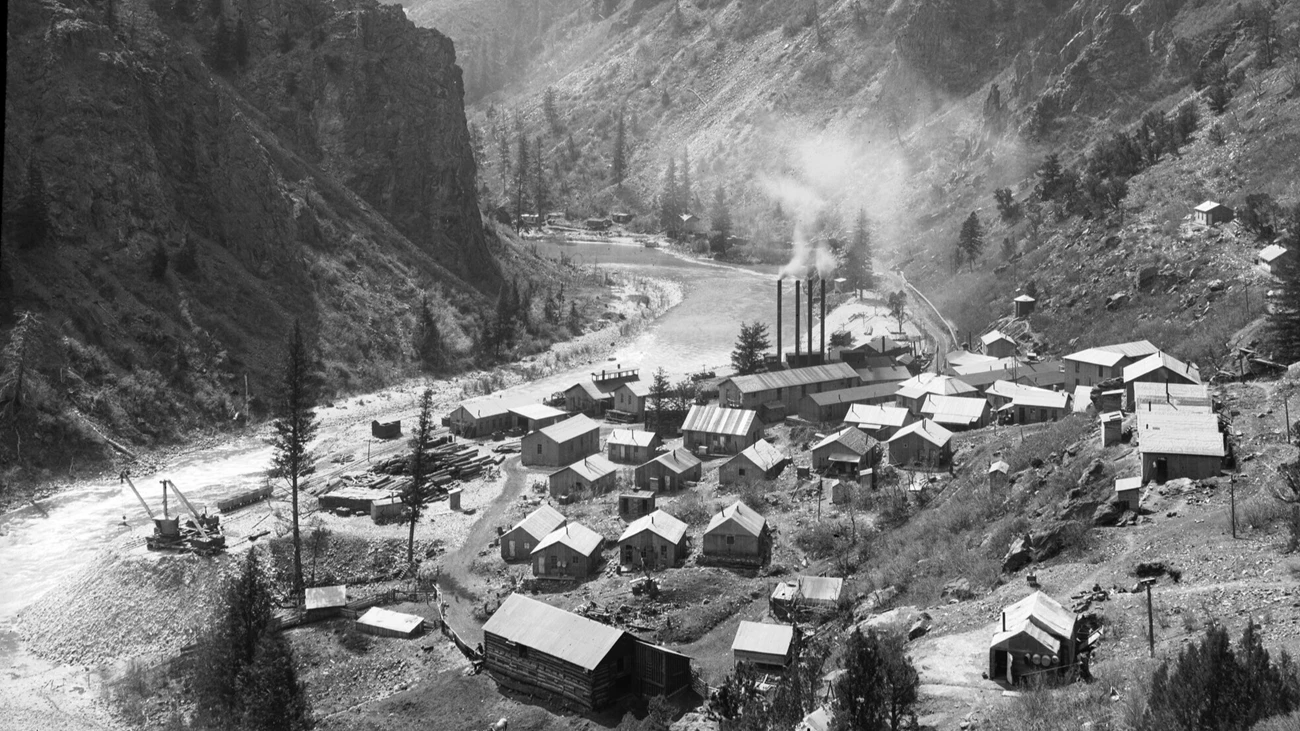 East Portal and Gunnison River Historic image of the Gunnison River and East Portal. Workshops, a powerhouse, and other frame buildings are near ore cars, piles of timber, and railroad tracks. A river between canyon walls runs by the town.