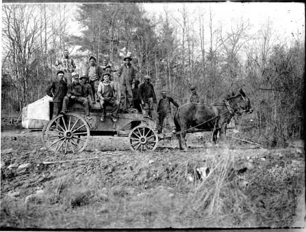 A historic black and white image of a wooden wagon, filled with men in work clothes, pulled by a team of two mules.
