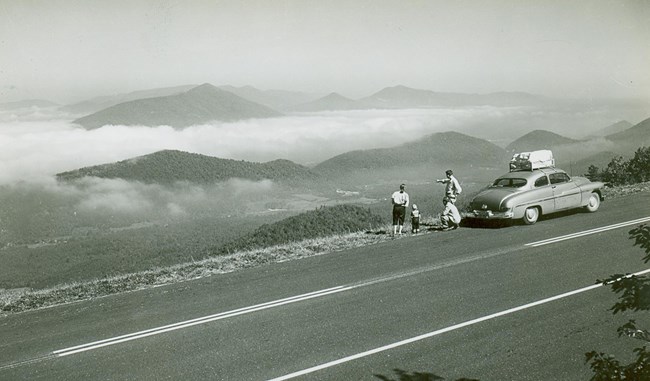 A family in a mid-1900s automobile stopped at an overlook to enjoy the scenery