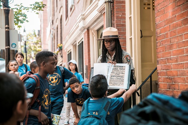 A park ranger holds a binder with a fugitive slave broadside as students look and listen
