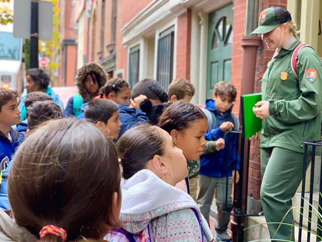 A park ranger interacting with young students on a trip. Ranger stands on front steps of a historic home.
