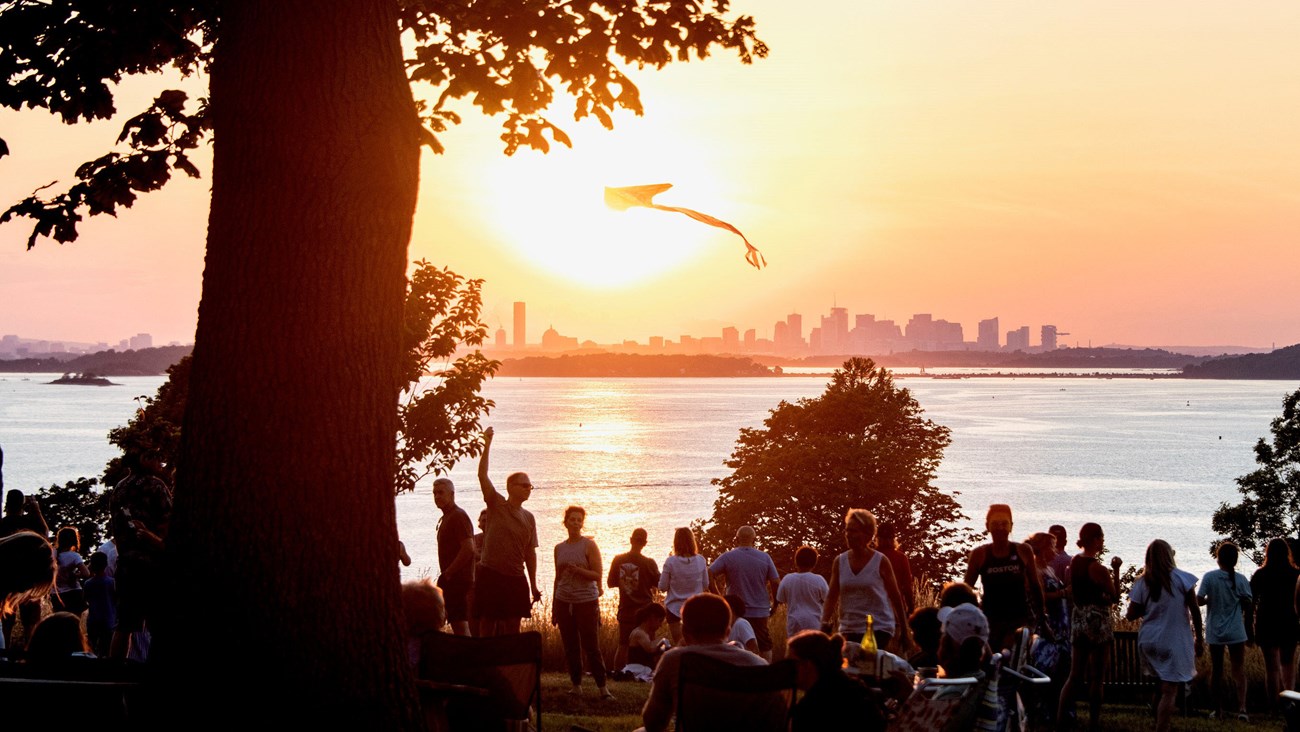 Group of people relaxing on an overlook of the harbor at sunset. A kite flies above them.