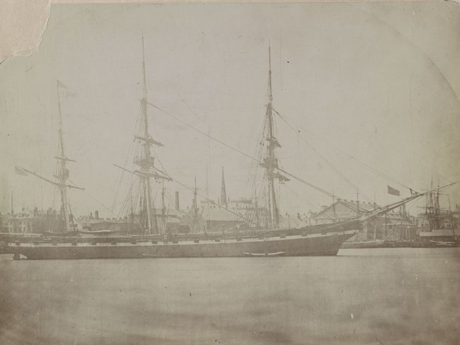 Photograph of USS Hartford with her masts fitted out. She is in a harbor with a waterfront area in th background.