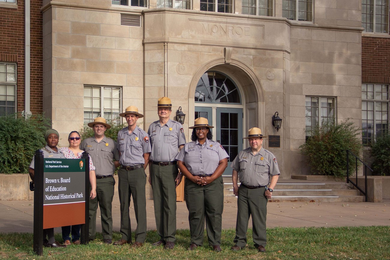 Park rangers stand behind Brown v. Board sign in front of brick building.