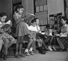 African American girl standing and reading with other children and teacher in the background.